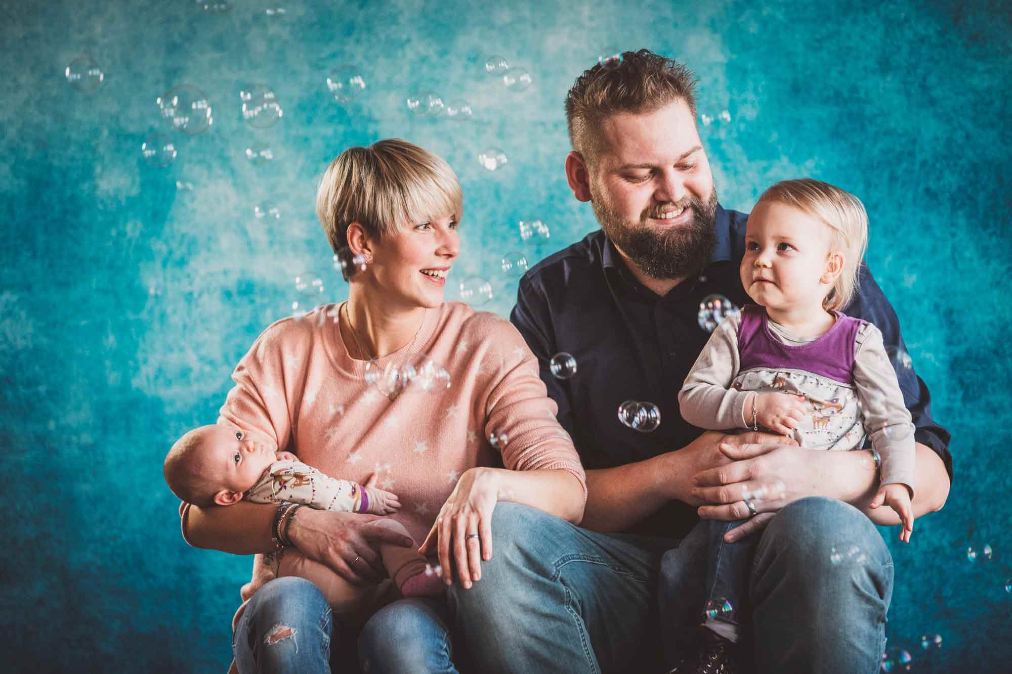 Familie, Eltern mit Kleinkind und Neugeborenem auf den Armen, mit Seifenblasen, vor blauem Hintergrund
