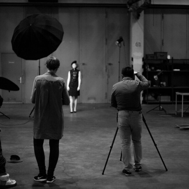 Fashionshooting, Fotograf mit Rücken zur Kamera fotografiert Model im Hintergrund, daneben steht ein Assistent, Making-of, Schwarz-Weiß