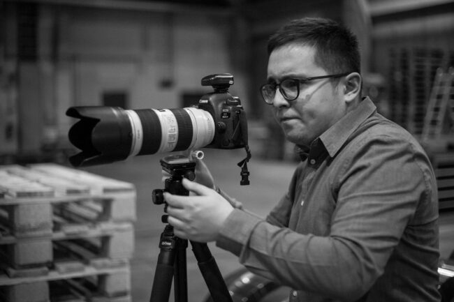 Fashionshooting, Fotograf mit Kamera auf Stativ vor industriellem Hintergrund, Seitenansicht, Schwarz-Weiß