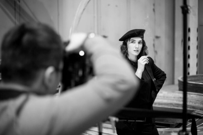 Fashionshooting, Fotograf steht mit Rücken zur Kamera und fotografiert Model in Uniform vor industriellem Hintergrund, Schwarz-Weiß