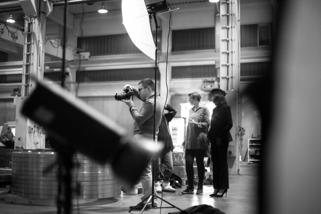 Fashionshooting, Fotograf schaut auf Kamera, daneben steht ein Model in Uniform und eine weitere Frau, vor industriellem Hintergrund, Lichtaufbau des Shooting ist zu sehen, Schwarz-Weiß