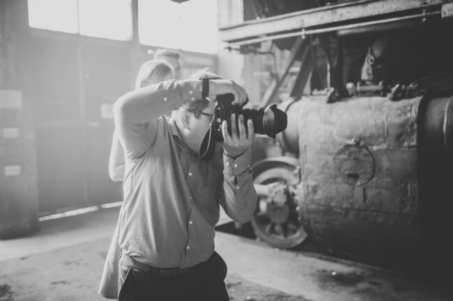 Fashionshooting, Fotograf mit Kamera in den Händen schießt ein Foto, im Hintergrund teile einer alten Lokomotive und Personen, Schwarz-Weiß, Querformat