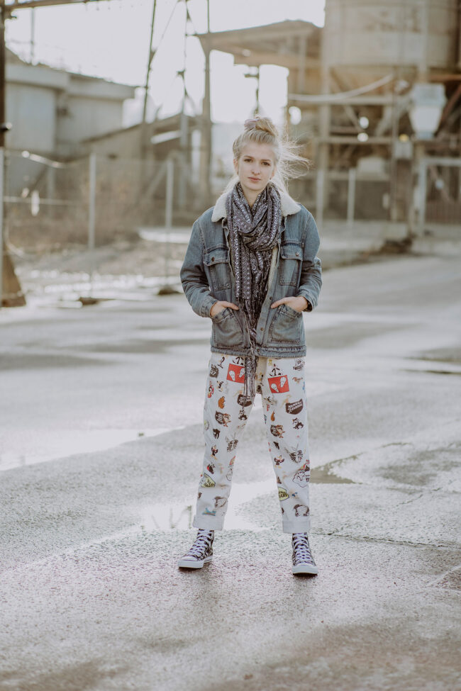 Portrait, junge Frau mit gemustertem Schal und gemusterter Hose an in Industriegebiet stehend, Hände in den Jackentaschen, Ganzaufnahme