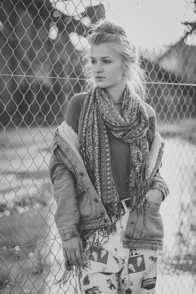 Portrait, junge Frau mit gemustertem Schal und gemusterter Hose an Maschendrahtzaun gelehnt, Halbportrait, Schwarz-Weiß