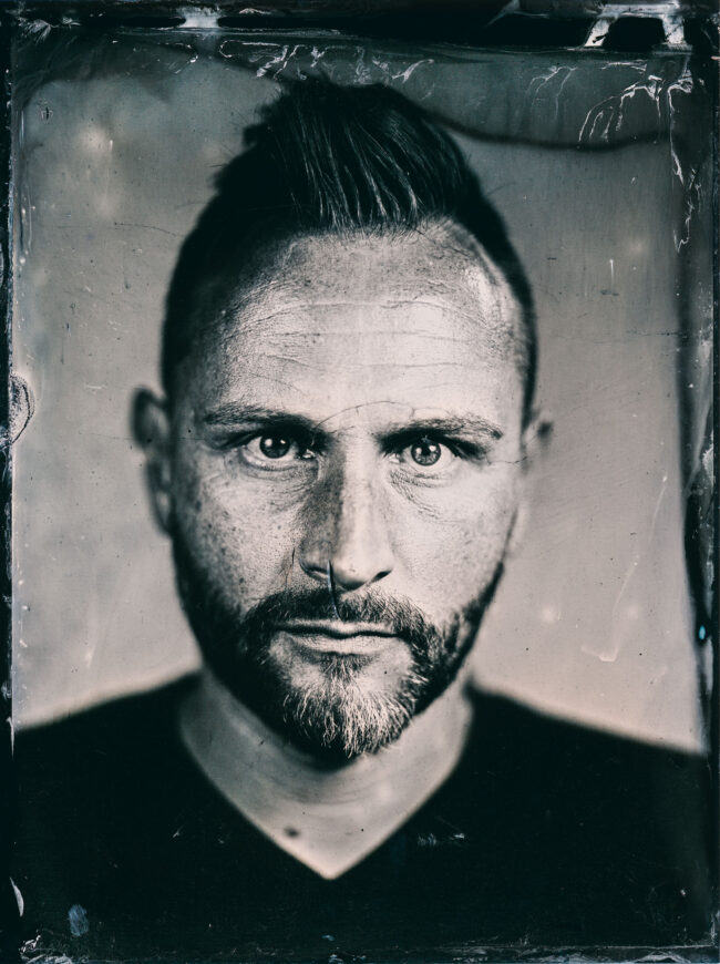 Nassplatte, Schwarz-Weiß, Portraitaufnahme von jungen Mann mit Bart und kurzer Irokesenfrisur auf hellem Hintergrund