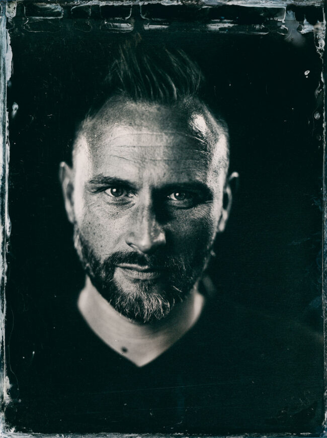 Nassplatte, Schwarz-Weiß, Portraitaufnahme von jungen Mann mit Bart und kurzer Irokesenfrisur auf schwarzem Hintergrund