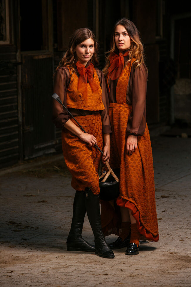 Fashion, zwei Models in orangenen Kostümen mit schwarzen Schuhen, einem Reithelm und einer Gerte auf gepflastertem Platz vor Stall stehend, Ganzaufnahme