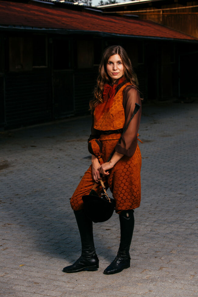 Fashion, Model in orangenem Kostüm mit schwarzen Reitstiefeln, einem Reithelm und einer Gerte auf gepflastertem Platz stehend, Ganzaufnahme