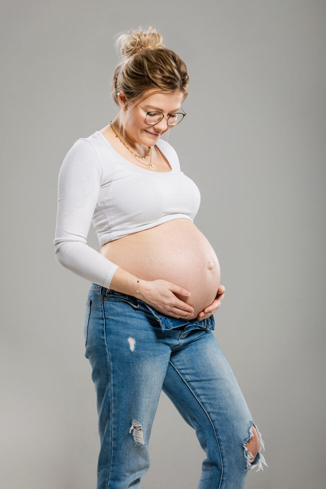 Babybauch, Frau in weißem Shirt und blauer Jeans hat Hände auf nackten Babybauch gelegt, Studioaufnahme vor grauem Hintergrund