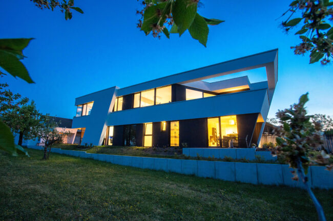 Architekturaufnahme, modernes zweistöckiges weißes Haus zur blauen Stunde, mit leicht abgeschrägten Vertikallinien, erleuchtete Fenster