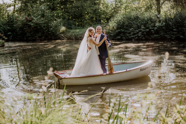 Hochzeit, Outdoorportrait, Brautpaar steht in Ruderboot auf einem See und lacht in die Kamera