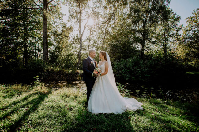 Hochzeit, Brautpaar schaut sich an, Brautstrauß gemeinsam haltend, an kleinem Bach im Wald, Querformat