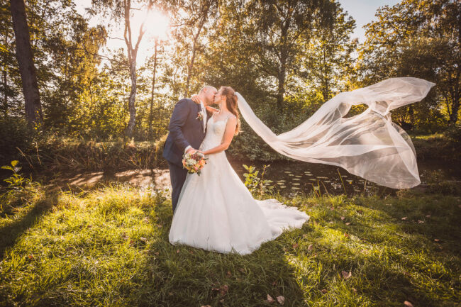 Hochzeit, Brautpaar sich küssend, mit wehendem Schleier, Brautstrauß gemeinsam haltend, an kleinem Waldteich, Querformat