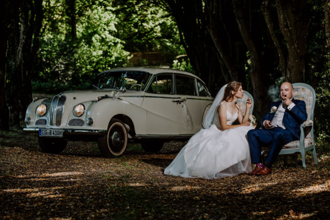 Hochzeit, Brautpaar sitz auf Stühlen vor Oldtimer im Wald, Bräutigam raucht Pfeife