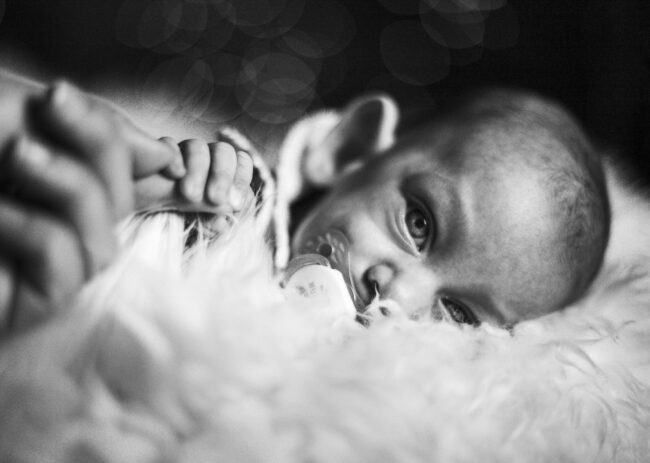 Baby, liegt seitlich auf einem Fell mit Schnuller im Mund, greif mit Hand den Finger der Mutter, schaut in die Kamera, Schwarz-Weiß