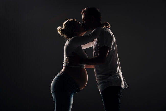 Babybauch, Mann und schwangere Frau stehen sich gegenüber, haben die Arme umeinander gelegt und küssen sich, Studioaufnahme mit Kantenlicht vor schwarzem Hintergrund, Halbaufnahme