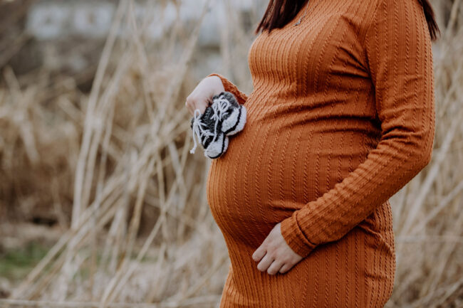 Babybauch, Outdoor, schwangere Frau in orangenem Kleid steht zwischen hohen Gräsern und hält kleine Babyschuhe an den Bauch, Detailaufnahme Bauch