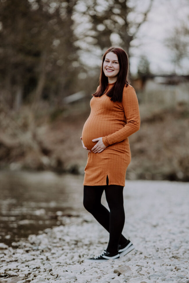 Babybauch, Outdoor, schwangere Frau in orangenem Kleid steht an Flussufer, hat beide Hände an den Bauch gelegt und strahlt in die Kamera
