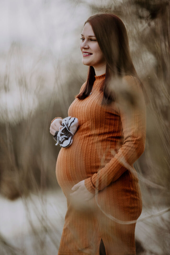 Babybauch, Outdoor, schwangere Frau in orangenem Kleid steht zwischen hohen Gräsern, hat beide Hände an den Bauch gelegt und hat kleine Babyschuhe in der Hand, Halbaufnahme