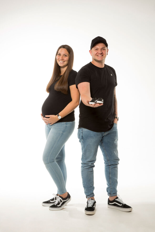 Babybauch, Mann und schwangere Frau stehen Schulter an Schulter, Frau hat Hände an Bauch gelegt, Mann hat Babyschuhe in der Hand, beide strahlen in die Kamera