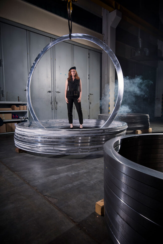 Fashion, Model in schwarzer Uniform und Barrett in Metallringen stehend vor industriellem Hintergrund, Hochformat, Ganzaufnahme