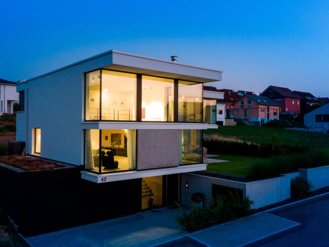 Architektur, Aufnahme modernes Haus in der blauen Stunde, durch große Fensterfronten sieht man Wohnbereich, Wohngebiet als Umgebung
