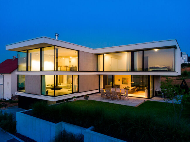 Architektur, modernes Haus über Eck mit großen Fensterfronten und Terrasse vor grünem Garten, Aufnahme zur blauen Stunde