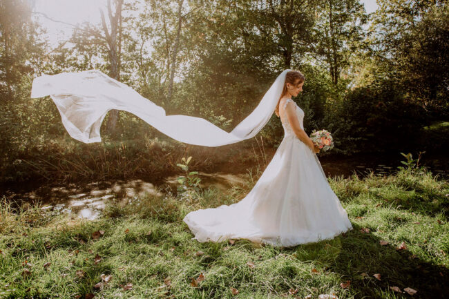 Hochzeit, Braut mit wehendem Schleier, auf Brautstrauß schauend in der Abendsonne, Querformat