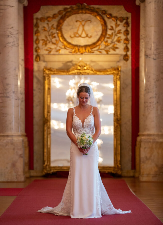 Hochzeit, Portrait, Braut steht in Marmorsaal vor einem Spiegel, auf einem roten Teppich und schaut auf Strauß, Ganzbild