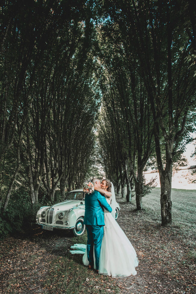 Hochzeit, Brautpaar küsst sich und steht vor Oldtimer in einer Allee
