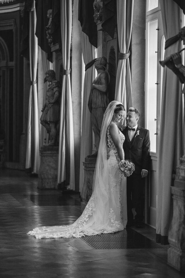 Hochzeit, Portrait, Ganzaufnahme von sich umarmenden Paar an einem Fenster in einem Schlafsaal. Bräutigam schaut Braut an, Braut schaut auf Blumen, schwarz-weiß