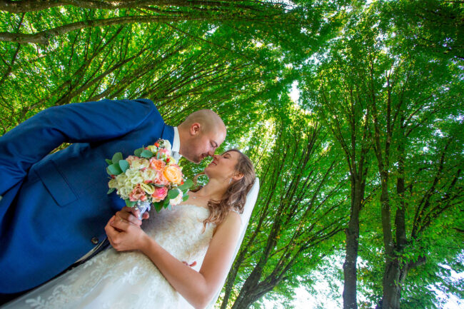 Hochzeit, Brautpaar steht Nase an Nase unter einem grünen Blätterdach, Halbaufnahme