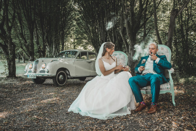 Hochzeit, Brautpaar sitz auf Stühlen vor Oldtimer im Wald, Bräutigam raucht Pfeife, Braut schaut man an und hält eine Kiste in der Hand