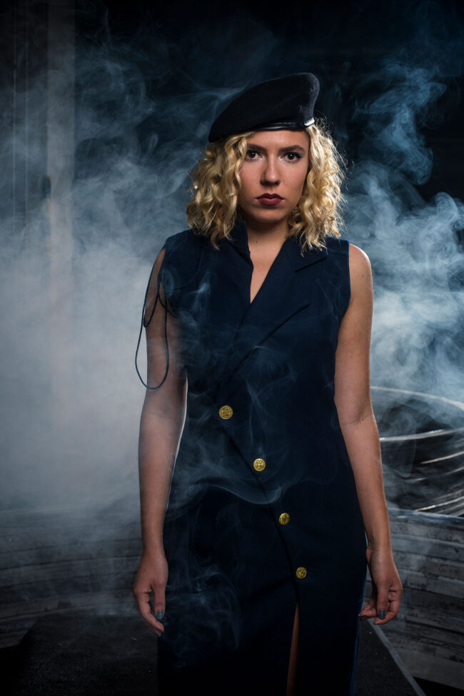 Fashion, Mode, junge Frau in dunkelblauem Kleid mit Goldknöpfen und hohem Beinschlitz, mit Barrett , vor dunklem Hintergrund im Nebel, Halbaufnahme