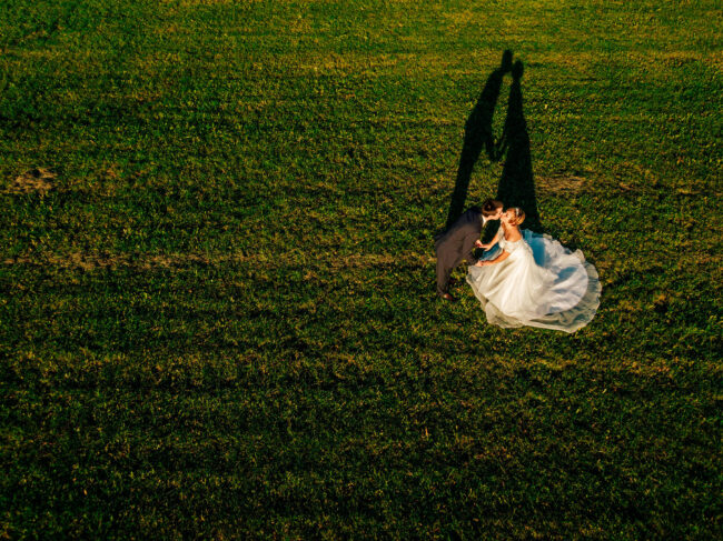 Hochzeit, Drohnenaufnahme, Brautpaar küsst sich auf einer Wiese in der Abendsonne, es ist ein langer Schatten des Paares zu sehen