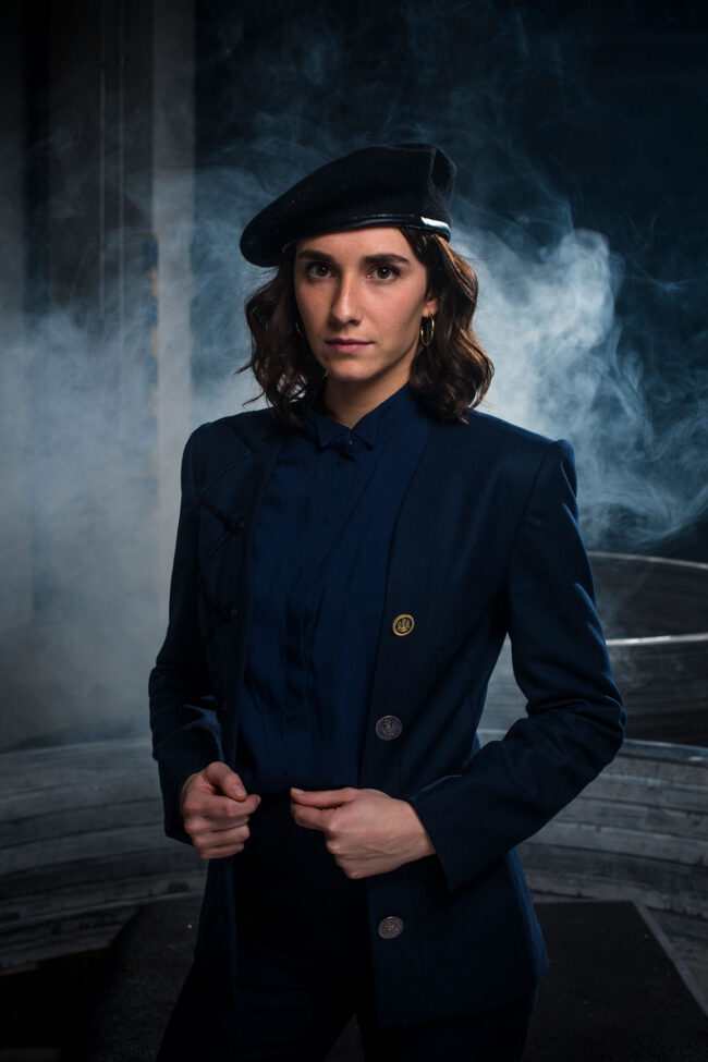 Fashion, Halbbildern von Frau mit dunkelblauer Bluse und Jacke mit goldverzierten Knöpfen und einem Hut, im Hintergrund ist Nebel