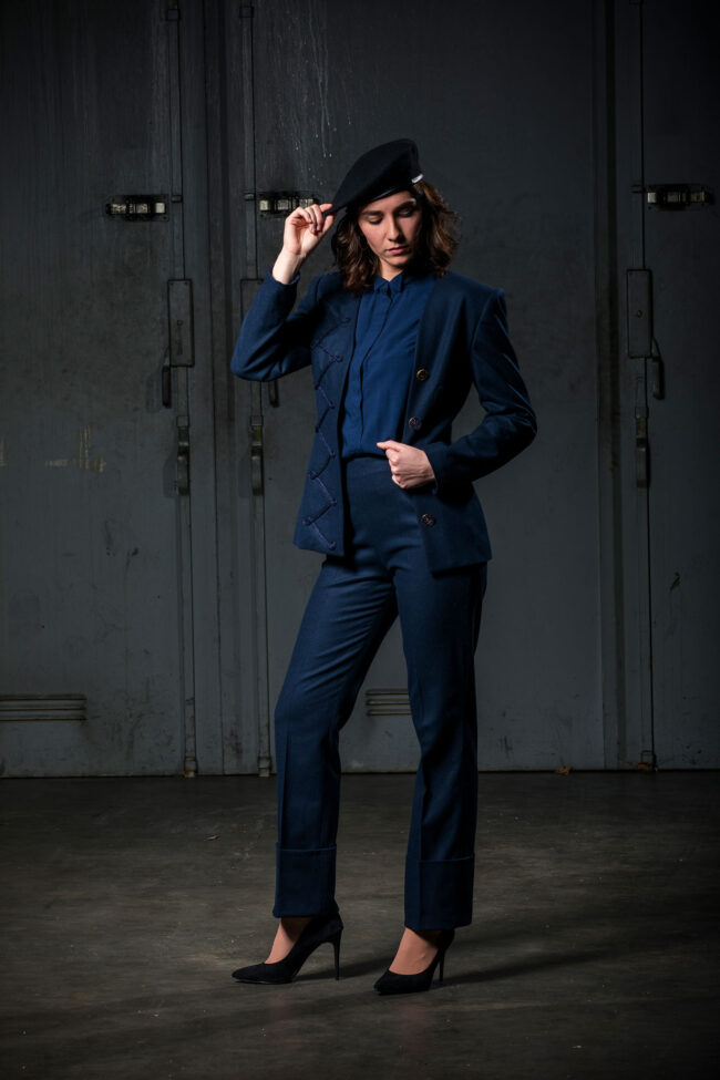 Fashion, Mode, junge Frau mit dunkelblauem Kostüm und Bluse, greift mit Hand an Hut, vor dunkelgrauen Metalltüren