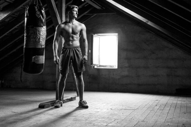 Portrait, Junger Mann neben Boxsack auf altem Dachboden, Ganzaufnahme, Schwarz-Weiß