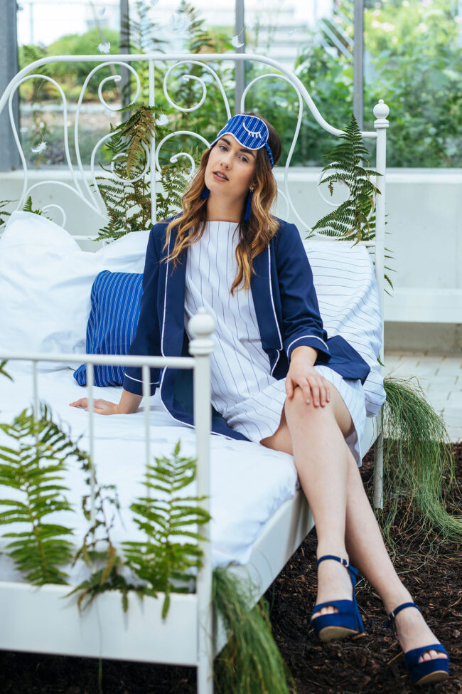 Fashion, Model in elegantem Nachthemd und dunkelblauem Gewand auf weißem Bett sitzend, in Gewächshaus, Ganzaufnahme