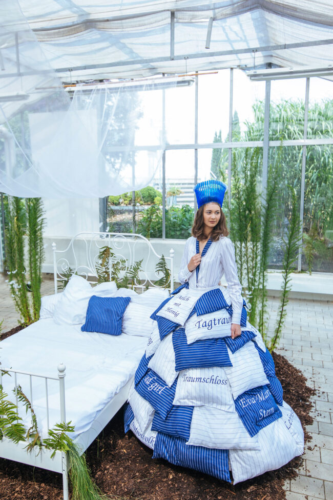 Model in Weiß-blauem Kleid aus Kissenbezügen vor einem weißen Bett in einem Gewächshaus