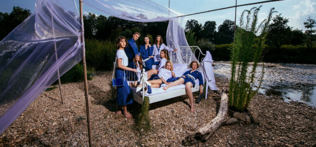 Gruppe an Models in Blau-weißer Kleidung auf und neben einem weißen Bett, das an Flussufer steht, Querformat