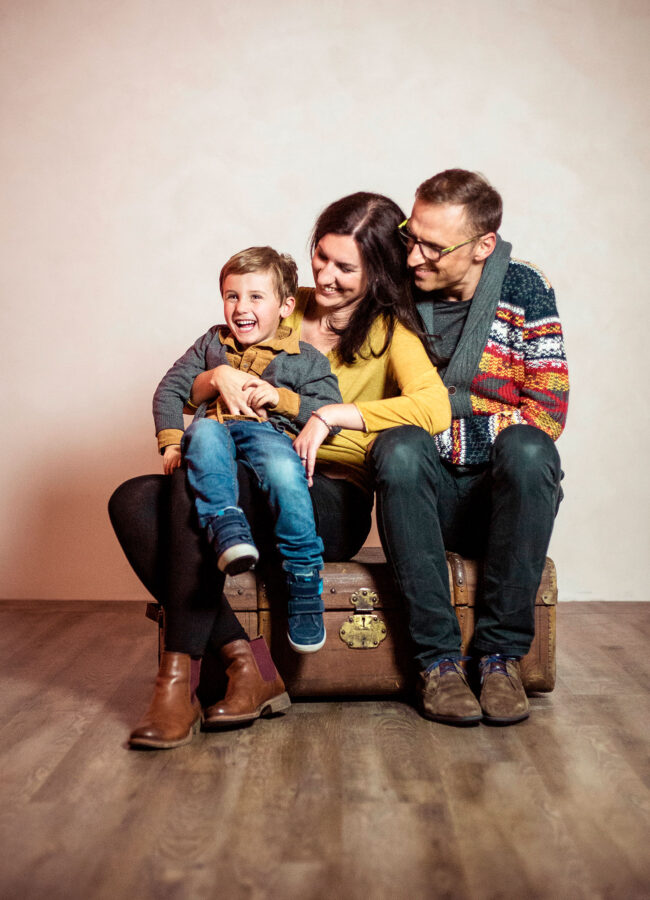 Familie, Eltern mit Kleinkind auf dem Schoß sitzen auf braunem Koffer, Eltern schauen zum Kind, Kind lacht in die Kamera