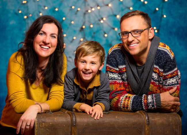 Familie, Eltern mit Kleinkind knien hinter braunem Koffer, vor blauem Hintergrund mit Lichterkette