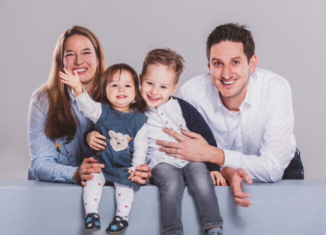 Familie, Eltern mit zwei Kindern, Kinder sitzen auf Bank, Eltern knien dahinter, strahlen in die Kamera, kleines Mädchen winkt, Studioaufnahme