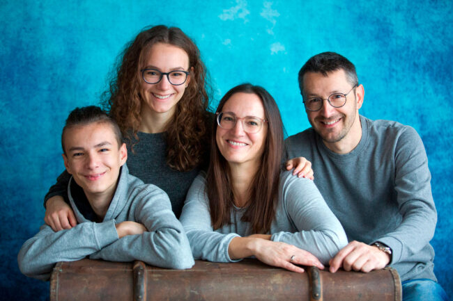 Familien, Eltern mit zwei Teenagern lehnen auf altem braunem Koffer vor blauem Hintergrund, alle strahlen in die Kamera