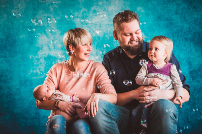 Familie, Eltern mit Kleinkind und Neugeborenem auf den Armen, mit Seifenblasen, vor blauem Hintergrund