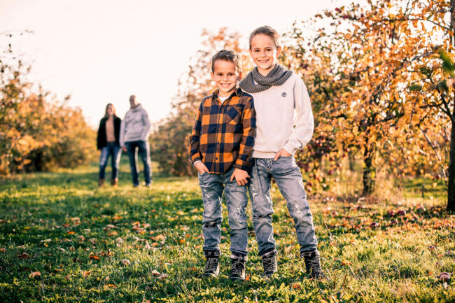 Familie, Outdoor, im Vordergrund zwei Jungs, die nebeneinander stehen und in die Kamera schauen, im unscharfen Hintergrund die Eltern Hand in Hand