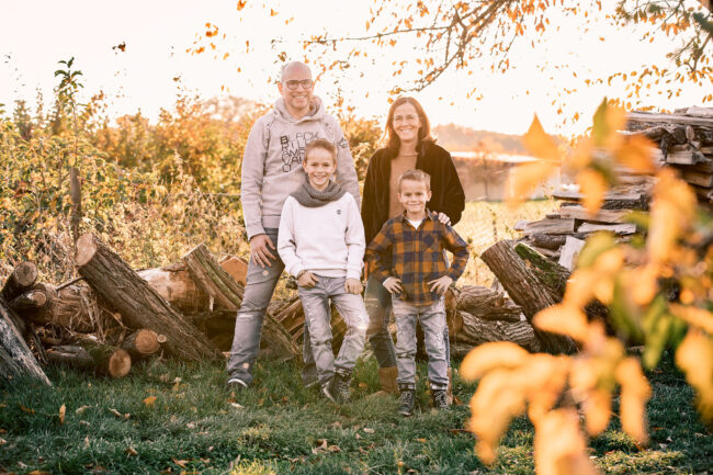 Familie,Outdoor, Eltern mit zwei Kindern stehen vor Holzstapel und lächeln in die Kamera