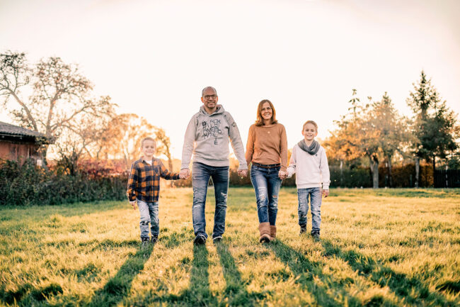 Familienshooting, Outdoor, Eltern mit zwei Kindern auf Wiese, laufen auf Kamera zu, halten sich an Händen, Sonnenuntergang