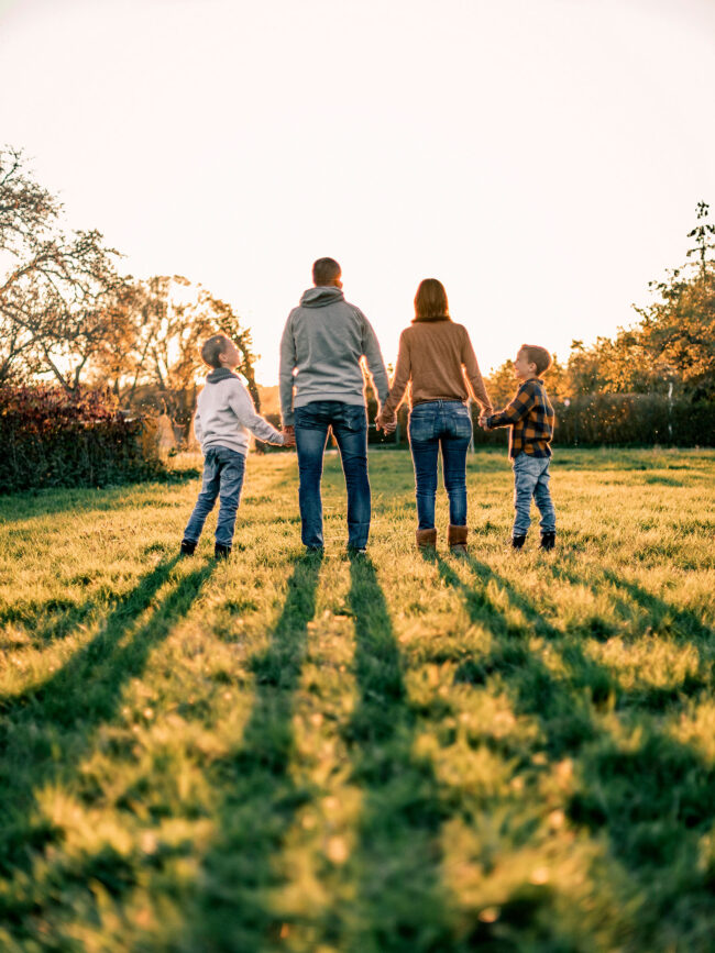 Familienshooting, Outdoor, Eltern mit zwei Kindern stehen auf Wiese mit Rücken zur Kamera, halten sich an Händen, Sonnenuntergang